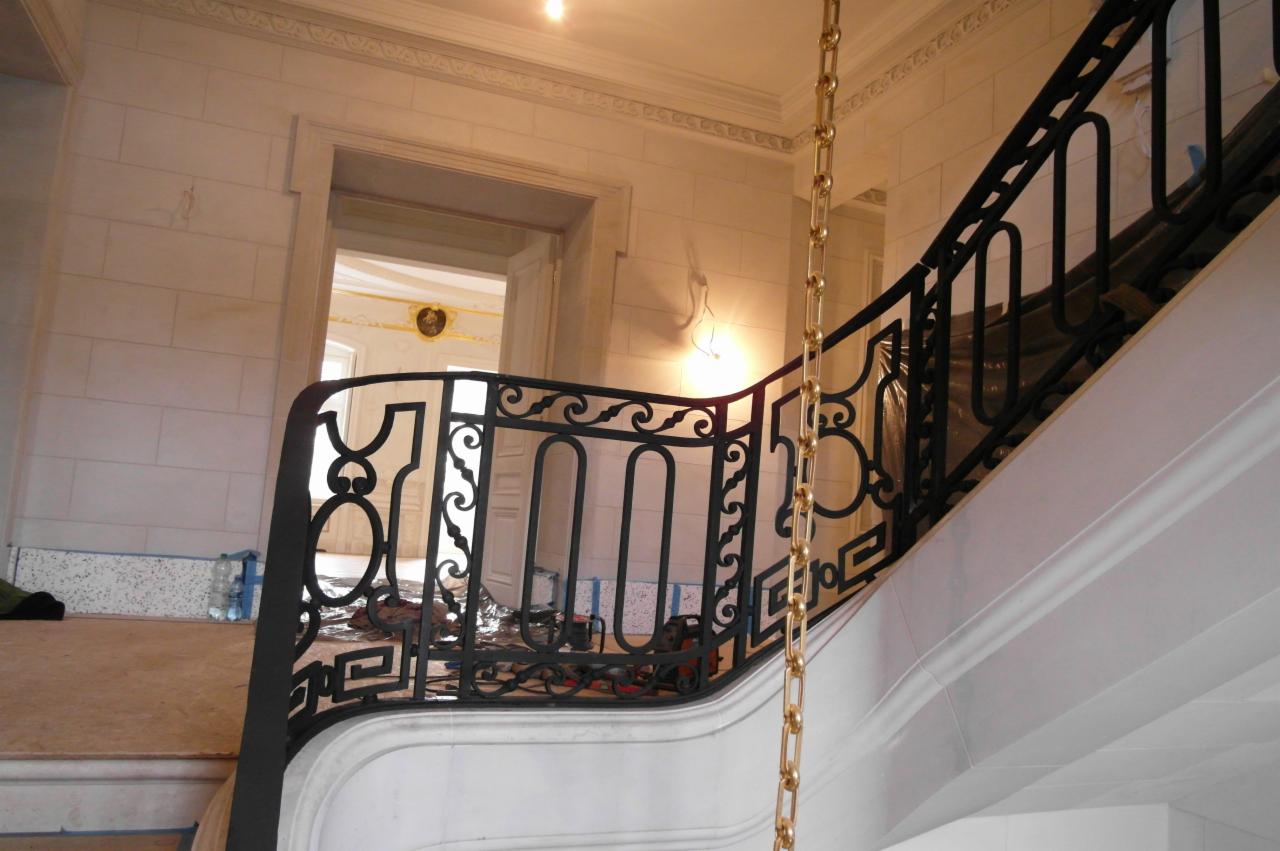 Klatka schodowa w trakcie remontu, widoczna czarna, ozdobna balustrada z metalu, elementy zabezpieczone folią, w tle otwarte drzwi do innego pomieszczenia.