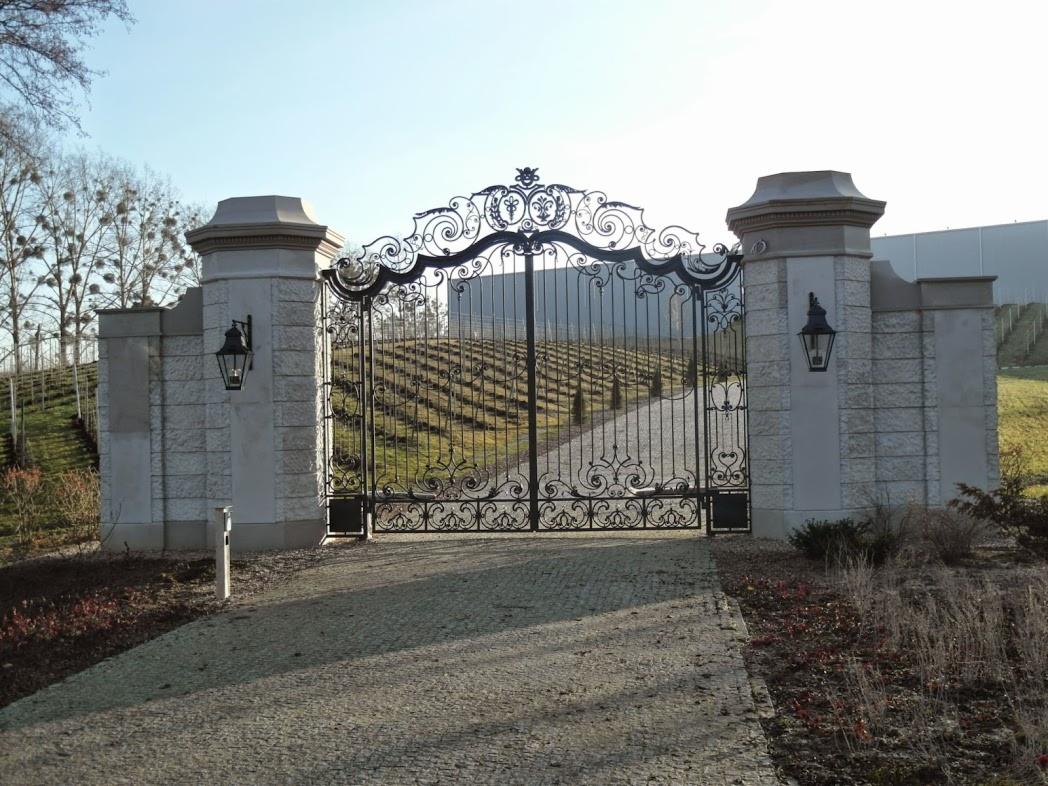 Kuta brama wjazdowa z ozdobnymi detalami, osadzona na kamiennych filarach z lampami. W tle widok na winnicę i budynek.