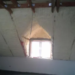 Poddasze po ociepleniu pianką PUR, widoczne drewniane belki stropowe i okno dachowe. Biała pianka szczelnie wypełnia przestrzeń między krokwiami.