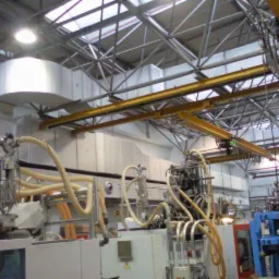 Instalacja przemysłowa z widocznymi przewodami i elementami wentylacji pod sufitem hali. Żółte rury i metalowa konstrukcja sufitu.