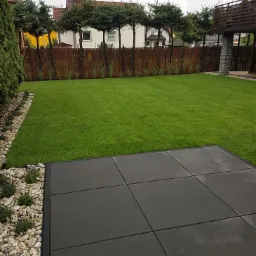 Ujęcie ogrodu z równo przystrzyżonym trawnikiem, ciemnymi płytami tarasowymi i obrzeżem z kamieni oraz niskich krzewów, oddzielającym trawnik od drewnianego płotu z nasadzeniami.