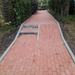 Czerwona ścieżka z kostki brukowej prowadząca przez ogród, z niskimi schodkami i szarymi obrzeżami, otoczona zielenią.