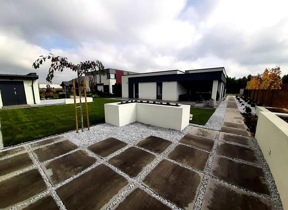 Nowoczesny ogród z geometrycznym układem ścieżki z płyt betonowych i białego żwiru, minimalistyczna architektura domu w tle, młode drzewko z brązowymi liśćmi.