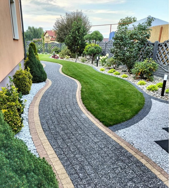 Kaskadowa ścieżka z kostki brukowej wijąca się przez zadbany trawnik, otoczona ozdobnymi krzewami i niskimi lampami ogrodowymi, z widokiem na dom w tle.