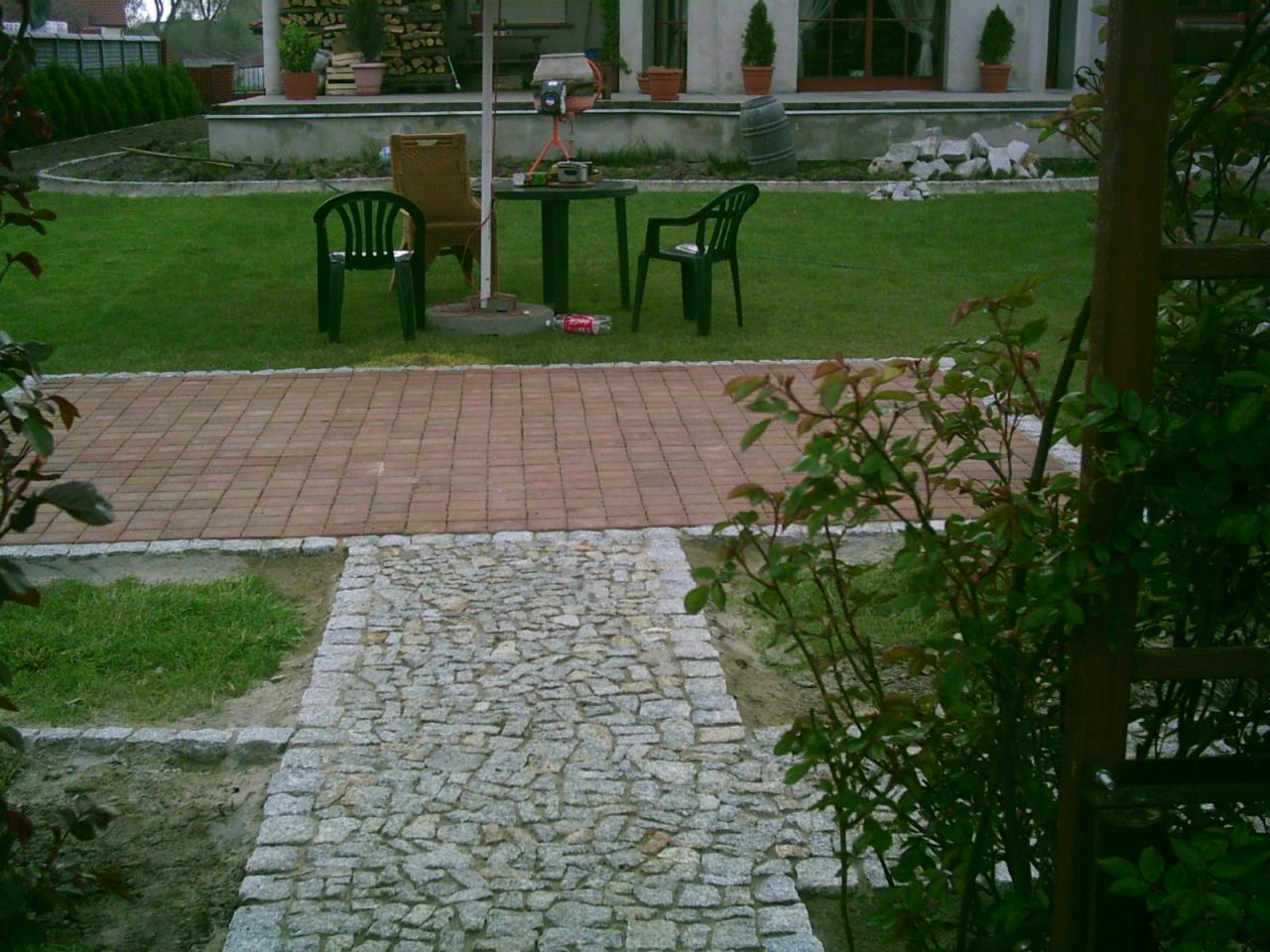 Ogród z alejką z kostki granitowej przechodzącą w czerwoną kostkę brukową. W tle trawnik, meble ogrodowe i roślinność.