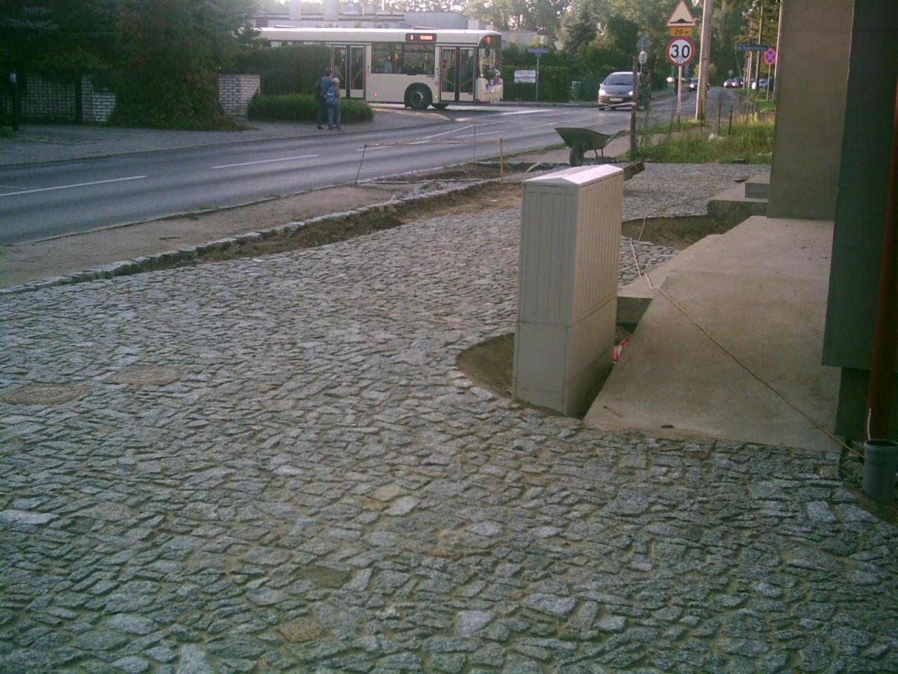 Ułożona kostka granitowa w odcieniach szarości przed budynkiem, z widocznymi elementami infrastruktury miejskiej w tle. Fragment chodnika w trakcie prac.