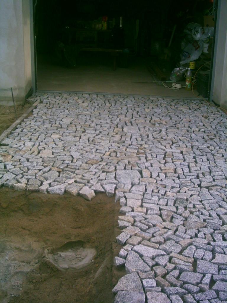 Ułożona kostka granitowa przed garażem, widoczny fragment wykopu pod dalszą część nawierzchni. Nieregularny kształt kamieni i szara kolorystyka.