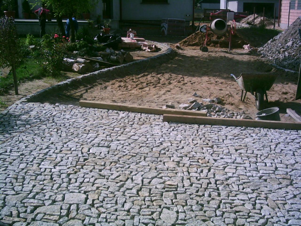 Ułożona kostka granitowa w ogrodzie, z fragmentem placu zabaw w budowie. Widoczna mieszarka i taczka, co sugeruje trwające prace wykończeniowe.
