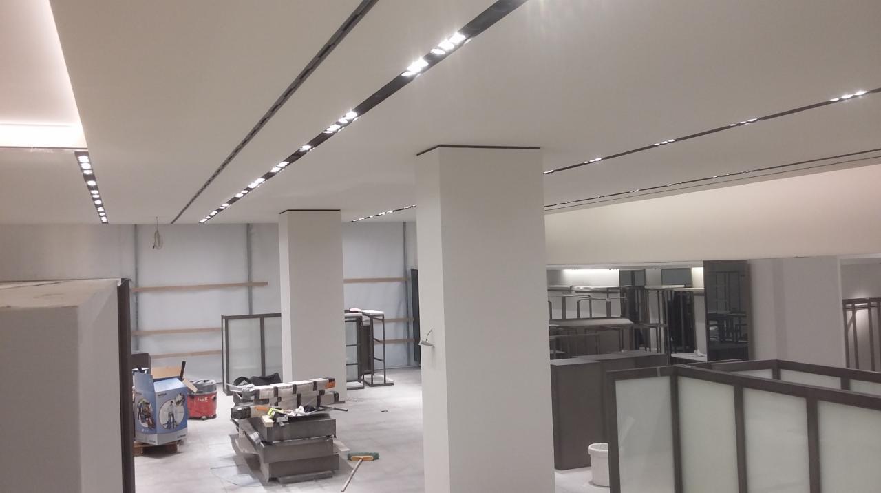 Jasne wnętrze sklepu w trakcie wykończenia, z białymi kolumnami i podwieszanym sufitem z liniowym oświetleniem. Widoczne elementy wyposażenia i narzędzia.