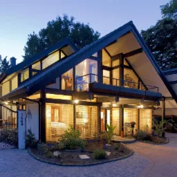 Dwukondygnacyjny dom z dużymi oknami i ciemnym dachem, oświetlony od wewnątrz, z zadbanym ogrodem i brukowanym podjazdem, widziany o zmierzchu.