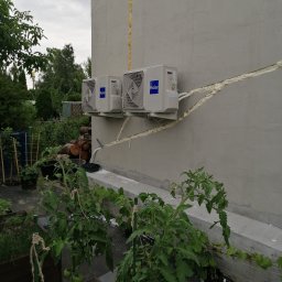 Euro-instal Piotr Ukleja - Dwa białe klimatyzatory Haier zamontowane na szarej ścianie budynku, widoczne przewody zabezpieczone pianką montażową, w tle zieleń ogrodu i uprawy pomidorów.