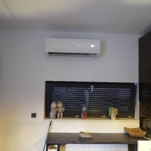 Biały klimatyzator ścienny zamontowany nad oknem z roletami w nowoczesnym wnętrzu, widoczne dekoracje na parapecie i fragment blatu z grzejnikiem.