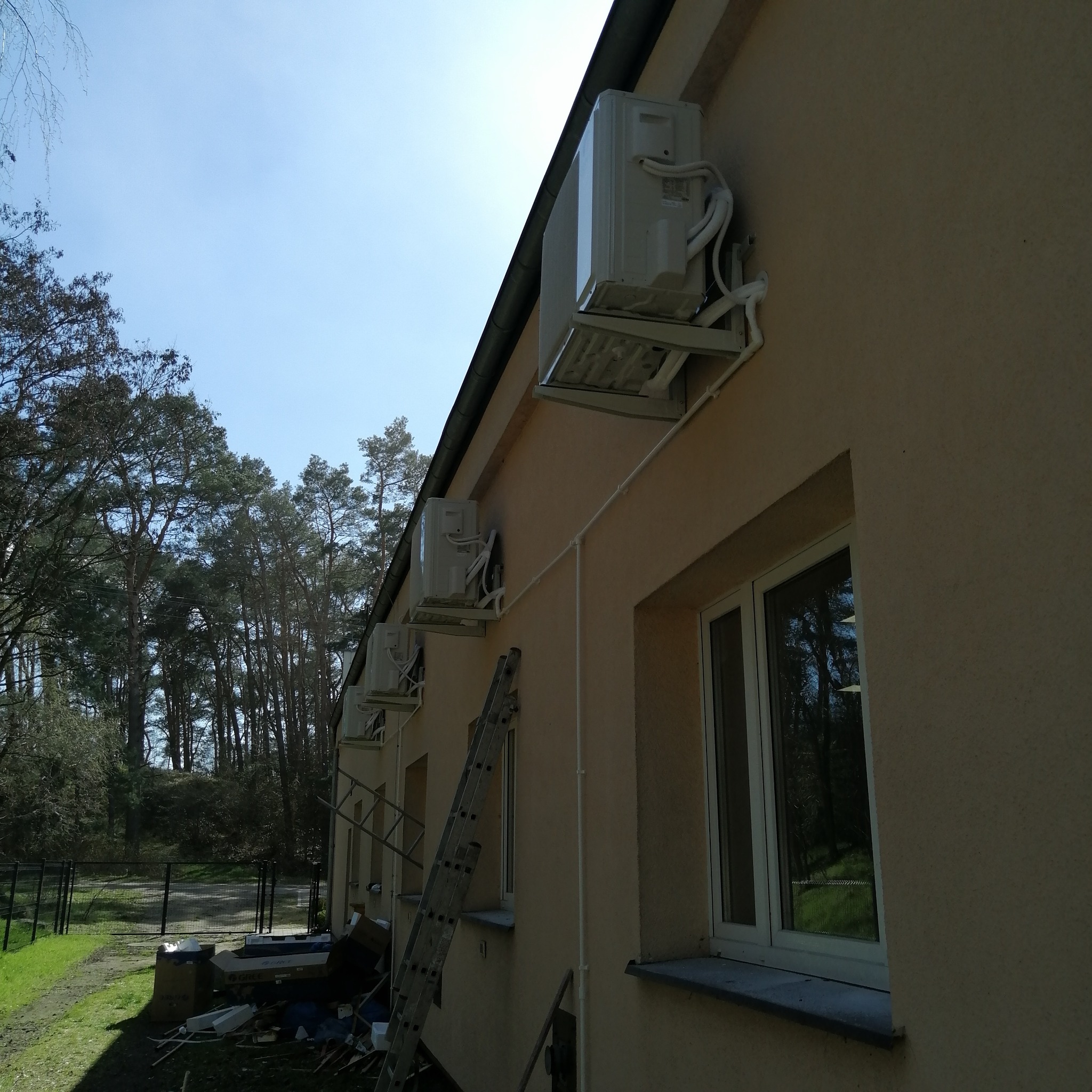 Zewnętrzne jednostki klimatyzacji zamontowane na elewacji budynku z widoczną drabiną i przewodami, tło stanowią drzewa i błękitne niebo.
