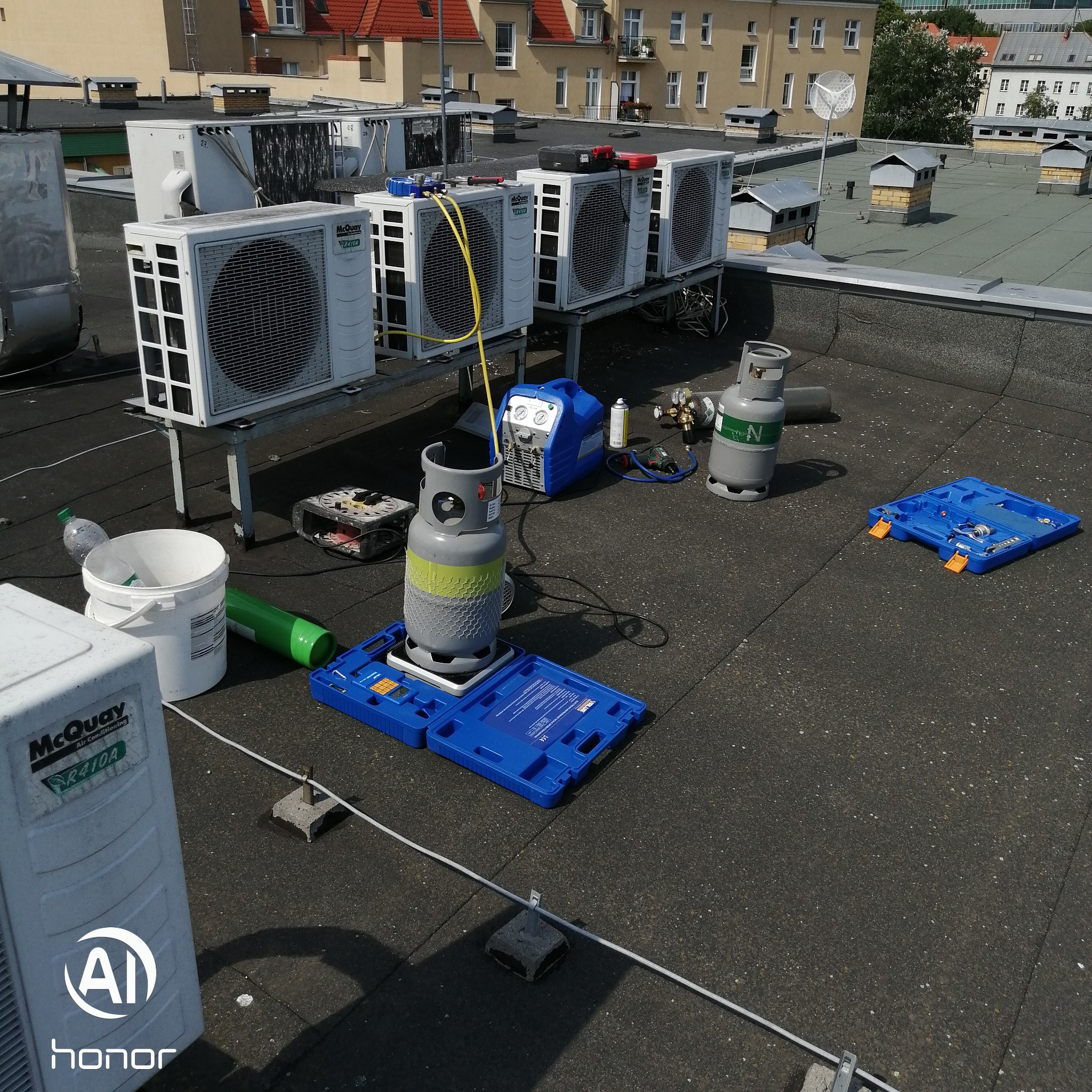 Serwis klimatyzacji na dachu budynku: widoczne jednostki zewnętrzne klimatyzatorów McQuay, butla z czynnikiem chłodniczym, narzędzia serwisowe, pompa próżniowa i przewody.