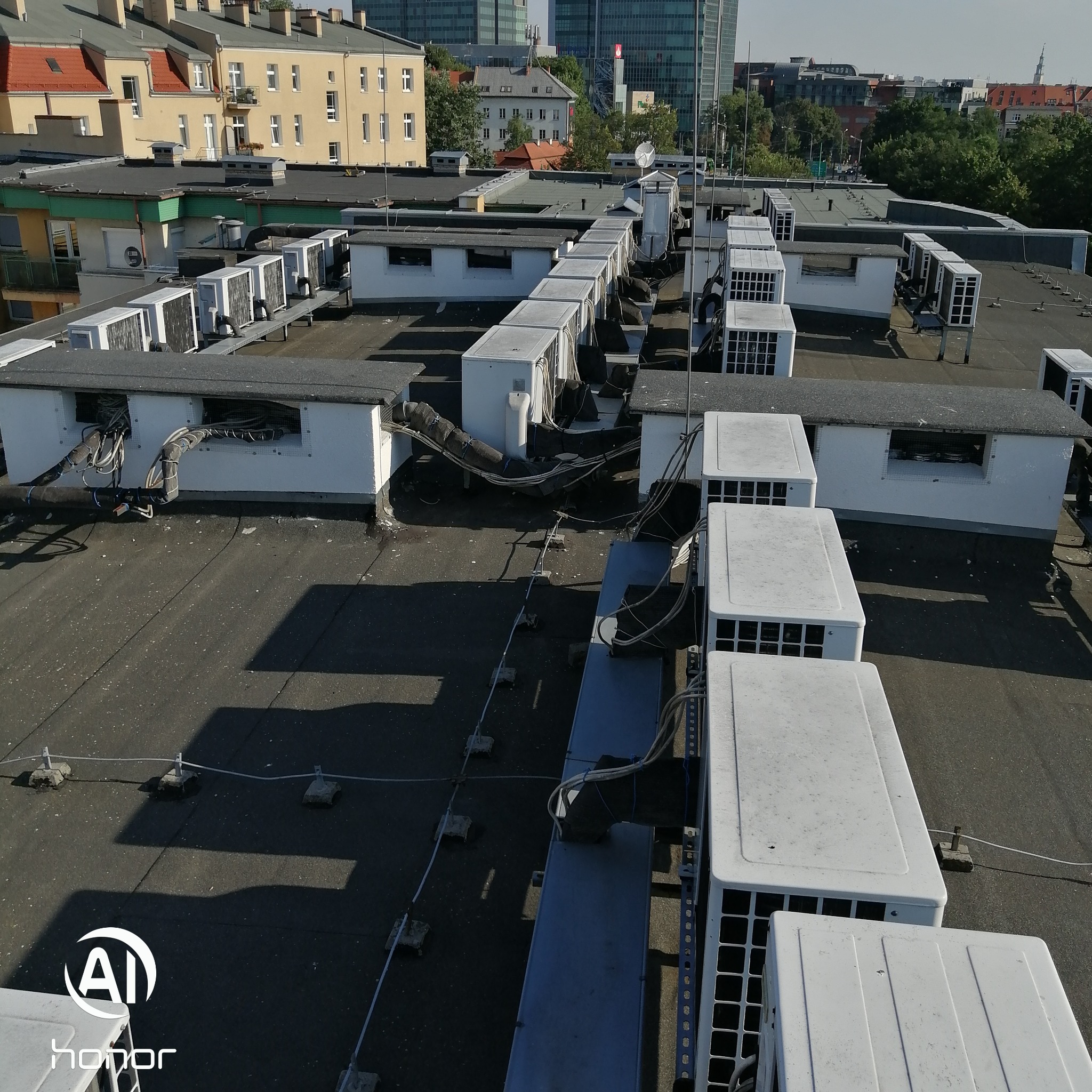 Widok z góry na liczne jednostki klimatyzacji i wentylacji na dachu budynku, z widocznymi przewodami i elementami mocującymi, na tle miejskiego krajobrazu.