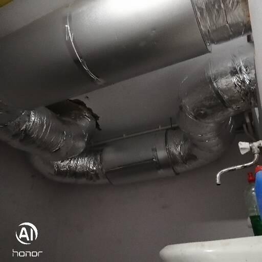 Srebrne, izolowane kanały wentylacyjne pod sufitem, z widoczną uszkodzoną izolacją i fragmentem instalacji wodnej obok.