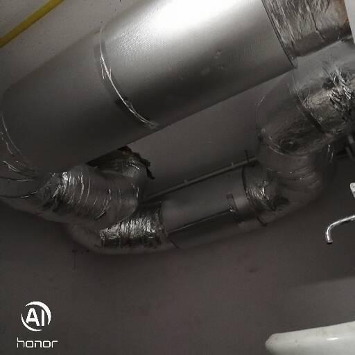 Izolowane kanały wentylacyjne pod sufitem, z widocznymi łączeniami taśmą aluminiową i fragmentem instalacji sanitarnej w tle.