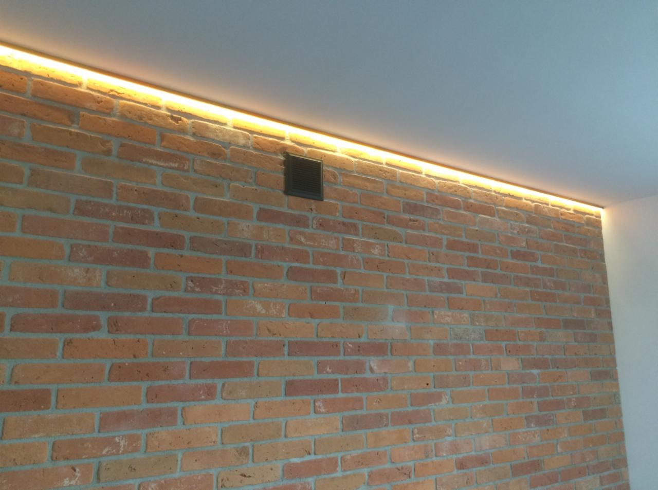 Ceglana ściana z dyskretnym oświetleniem LED podsufitowym i kratką wentylacyjną.