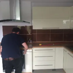 Montaż kuchni na wymiar: mężczyzna instaluje piekarnik w zabudowie z kremowych szafek z brązowymi płytkami. Poziomica i miarka leżą na blacie.