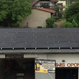 Panele fotowoltaiczne na dachu warsztatu samochodowego, widok z góry, w tle zabudowa jednorodzinna i drzewa.