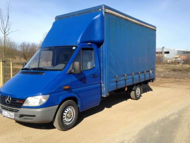 Niebieski bus Mercedes-Benz Sprinter z plandeką na tle błękitnego nieba i budynku. Widoczny numer rejestracyjny. Transport towarów.