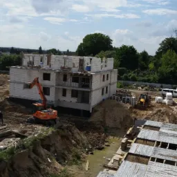 Budowa kompleksu mieszkalnego: dwupiętrowe budynki w stanie surowym, zbrojenia fundament&oacute;w, pomarańczowa koparka Hitachi i ż&oacute;łta ładowarka na placu budowy w słoneczny dzień.