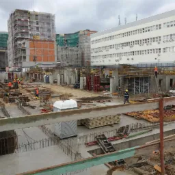 Szeroki plan budowy zbrojonych fundamentów pod budynek, widoczne szalunki, zbrojenia, betonowe słupy, pracownicy w kaskach i kamizelkach odblaskowych oraz budynki w tle.