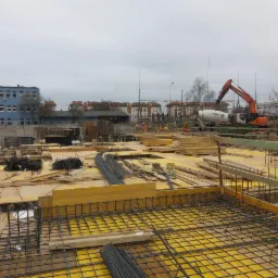 Żółte szalunki z siatką zbrojeniową na budowie, w tle betoniarka i koparka, widoczny zbrojarz przy pracy.