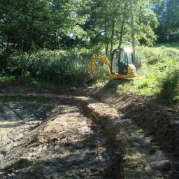 Żółta koparka gąsienicowa pracuje nad wykopem stawu w zarośniętym terenie; widoczny wykop z błotnistą wodą i świeżo naruszona ziemia.