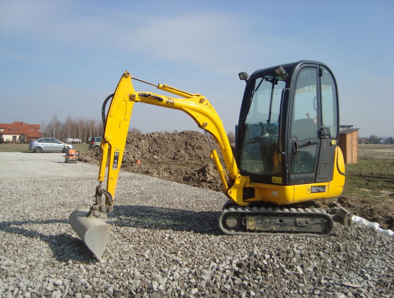 Żółta mini koparka JCB 8016 na żwirowym podłożu z wysięgnikiem i łyżką skierowanymi w lewo, w tle pryzma ziemi, domy i błękitne niebo.