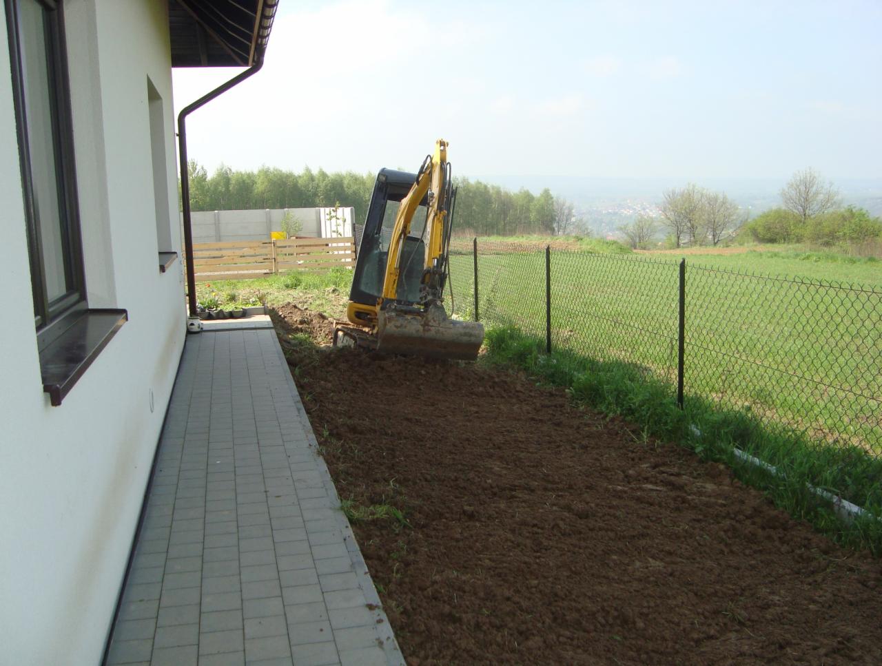 Żółta mini koparka przygotowuje teren przy budynku z szarą kostką brukową i metalową siatką ogrodzeniową w tle.