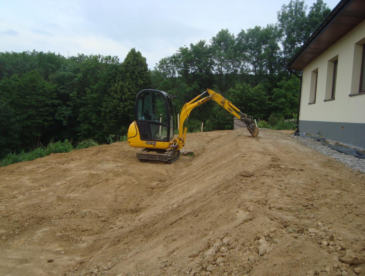 Żółta mini koparka na nierównym, piaszczystym terenie, w tle budynek z trzema oknami i zalesiony obszar pod pochmurnym niebem.