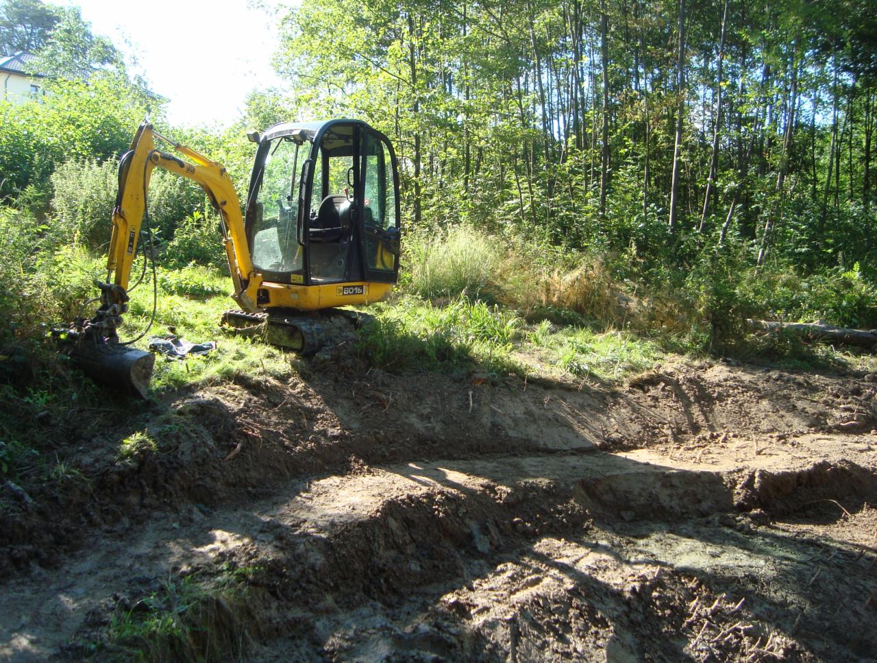 Żółta mini koparka JCB 8016 na terenie budowy, widoczne świeżo wykopane rowy w ziemi, zarośla i drzewa w tle.