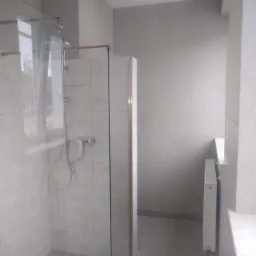 Nowoczesna kabina prysznicowa ze szklanymi ścianami i złotymi okuciami, zamontowana w łazience wykończonej jasnymi płytkami, widok na okno z zewnątrz.