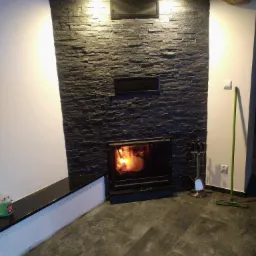 Wykończony kominek z czarnego łupka, płomień w palenisku, drewniane belki stropowe, ciemna podłoga z płytek, zestaw akcesoriów kominkowych, zielona zmiotka oparta o ścianę.