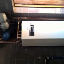 Biała jednostka grzewcza z podłączonymi miedzianymi rurami i zbiornikami, zamontowana na ciemnoszarej kafelkowej ścianie, widoczna w narożniku pomieszczenia.