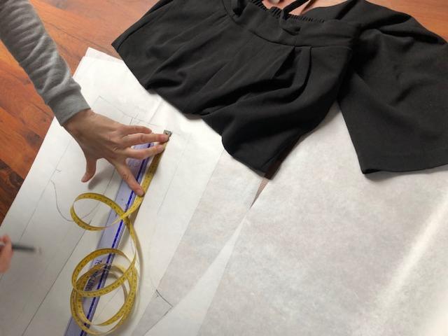 Ręka mierzy żółtą miarką wzór odzieży na papierze, obok leży czarna bluzka z szerokimi rękawami.