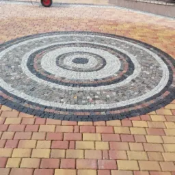 Okrągły wzór z kostki brukowej w odcieniach szarości, bieli i czerni na podjeździe z kolorowej kostki, z wózkiem i osobą w tle.