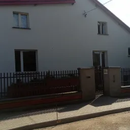 Jasnoszary dom jednorodzinny z czerwonym dachem i brązowym ogrodzeniem, widoczny z poziomu ulicy z brukowaną nawierzchnią i żwirowym poboczem.