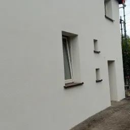 Świeżo otynkowana elewacja budynku z białym, strukturalnym tynkiem, widoczne okna z brązowymi parapetami i fragment rusztowania z boku.