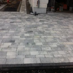 Ułożona szara kostka brukowa na posesji, widoczne obrzeża i przygotowane miejsce pod nasadzenia, w tle fragment budynku w trakcie budowy.