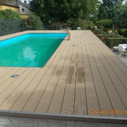 Beżowy taras kompozytowy otaczający basen z turkusową wodą, widoczny metalowy wodotrysk i oświetlenie wbudowane w deski, w tle zieleń ogrodu.