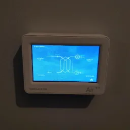 Ekran sterownika systemu wentylacji mechanicznej z odzyskiem ciepła, wyświetlający schemat przepływu powietrza, temperatury i wilgotność, zamontowany na szarej ścianie.