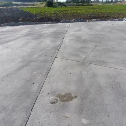 Świeżo wylana, szara powierzchnia z betonu jastrychowego z widocznymi szczelinami dylatacyjnymi, w tle pole uprawne i nasyp ziemi.