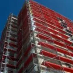 Budynek w rusztowaniach z czerwonymi osłonami siatkowymi, widok z dołu na błękitne niebo. Ocieplanie budynku, prace na wysokościach.