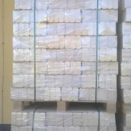 Ułożone na palecie i zabezpieczone folią pakiety brykietu drzewnego, widoczne zielone taśmy spinające, tło stanowi ściana budynku w kolorze żółtym i turkusowym.