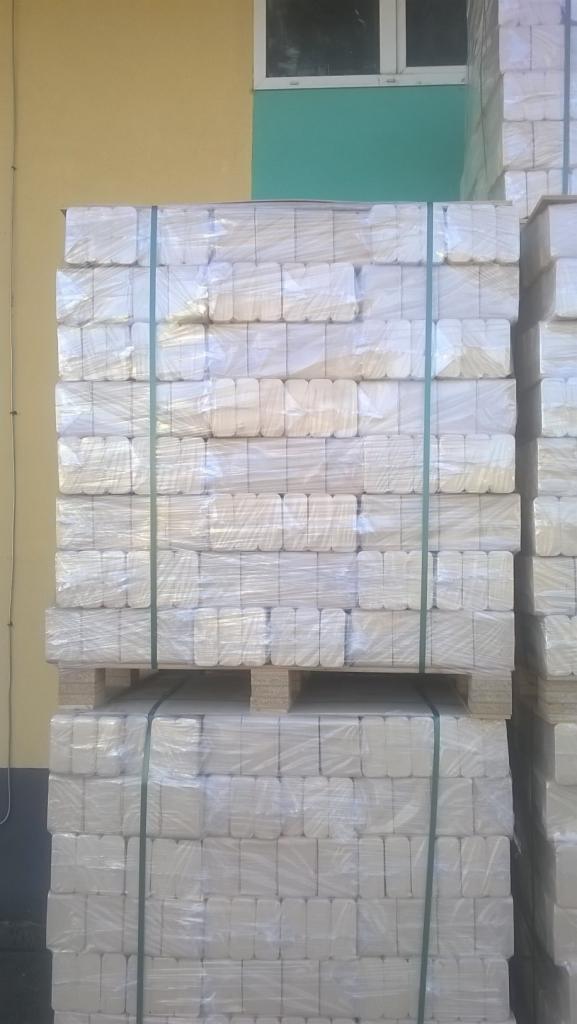 Ułożone na palecie i zabezpieczone folią pakiety brykietu drzewnego, widoczne zielone taśmy spinające, tło stanowi ściana budynku w kolorze żółtym i turkusowym.