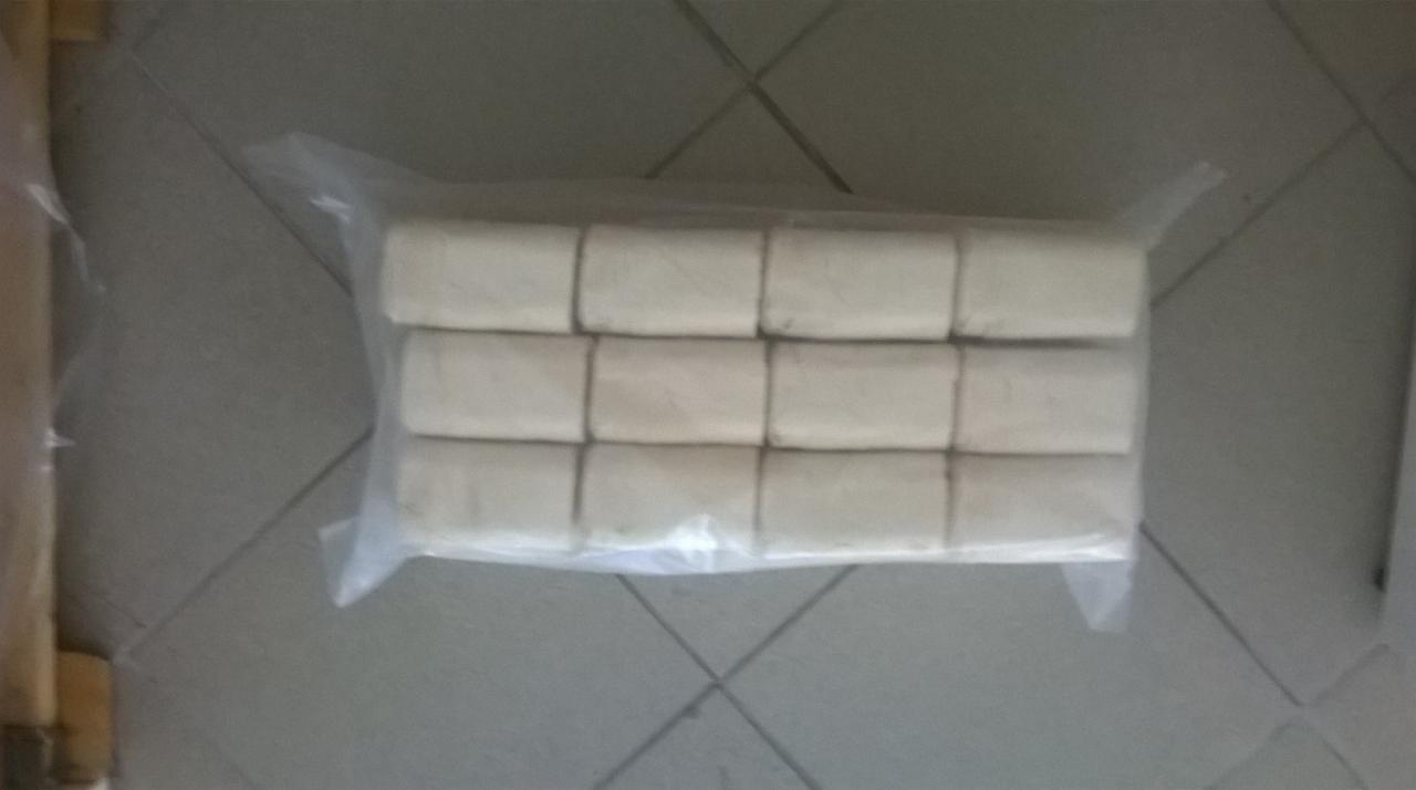 Kilkanaście sprasowanych kostek materiału, prawdopodobnie trocin lub wiórów, ułożonych w trzech rzędach i zapakowanych w przezroczystą folię na szarej podłodze z płytek.