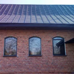 Ceglana ściana budynku z czterema łukowatymi oknami odbijającymi drzewa, z ciemnym, metalowym dachem z wyraźnymi liniami łączeń blach.