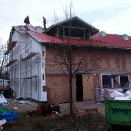 Budowa domu jednorodzinnego w trakcie, z widocznym rusztowaniem, częściowo ocieploną elewacją w kolorze białym, czerwoną blachodachówką na dachu i pracownikami na nim, oraz gruzem i odpadami...